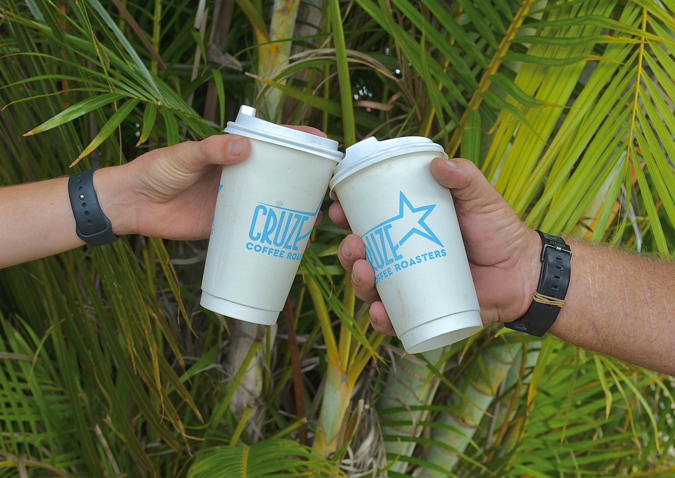 Two hands holding white coffee cups with visible branding against a green leafy background